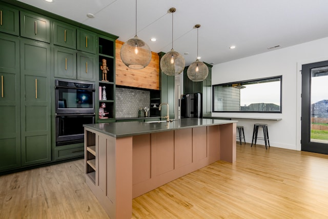 kitchen with a kitchen island with sink, double oven, stainless steel fridge, decorative backsplash, and green cabinetry