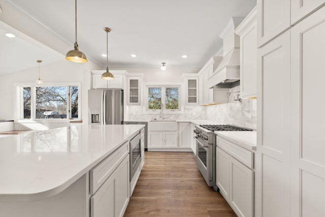 kitchen with white cabinetry, pendant lighting, appliances with stainless steel finishes, custom range hood, and hardwood / wood-style flooring