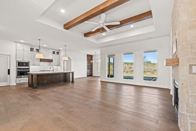 living room with sink, dark wood-type flooring, a stone fireplace, beamed ceiling, and ceiling fan with notable chandelier