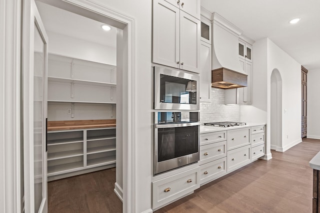 kitchen with decorative backsplash, custom range hood, stainless steel appliances, hardwood / wood-style floors, and white cabinetry