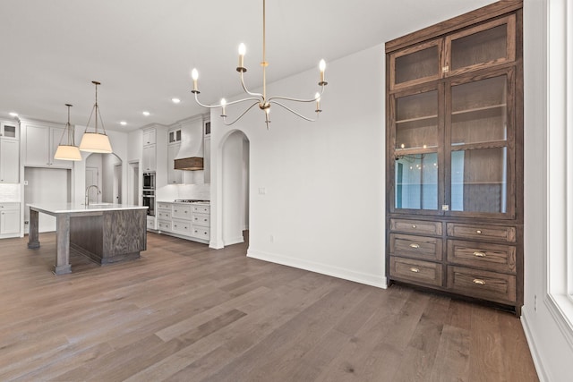 kitchen with premium range hood, sink, an island with sink, decorative light fixtures, and white cabinetry