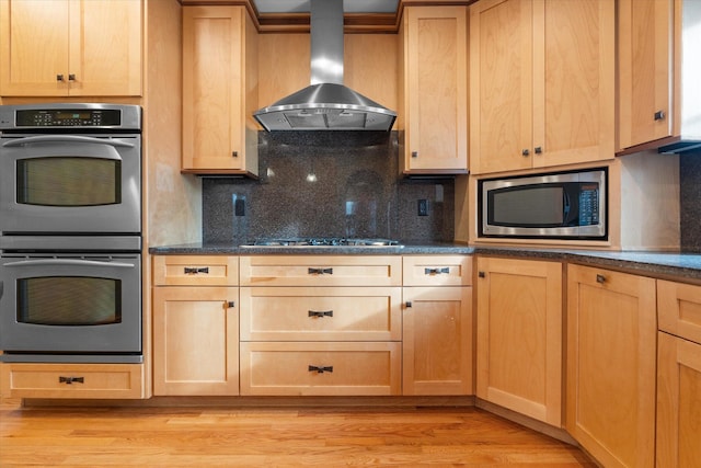 kitchen featuring wall chimney exhaust hood, dark stone countertops, tasteful backsplash, light hardwood / wood-style floors, and stainless steel appliances