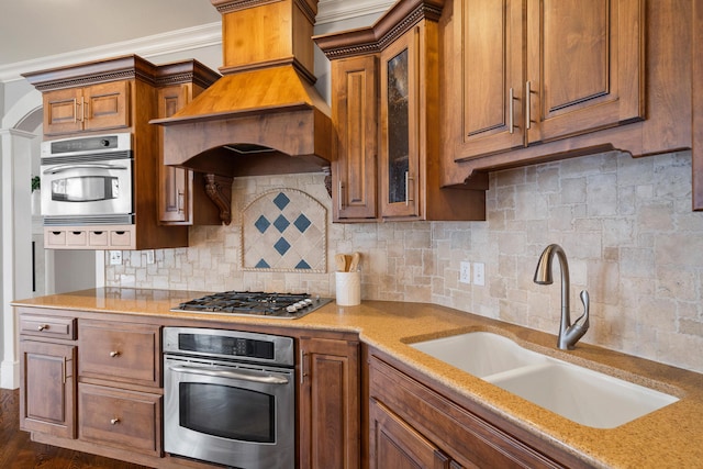 kitchen with stainless steel appliances, custom range hood, brown cabinetry, glass insert cabinets, and a sink