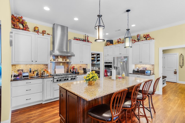 kitchen featuring a kitchen island with sink, white cabinets, wall chimney range hood, crown molding, and appliances with stainless steel finishes