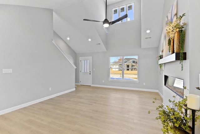 unfurnished living room featuring light wood finished floors, a high ceiling, a glass covered fireplace, ceiling fan, and baseboards