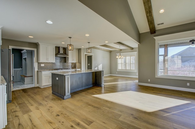 kitchen featuring wall chimney exhaust hood, white cabinetry, a large island with sink, stainless steel refrigerator, and pendant lighting