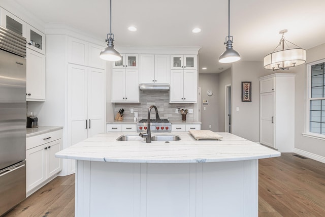 kitchen with built in fridge, decorative light fixtures, white cabinets, light stone counters, and a center island with sink