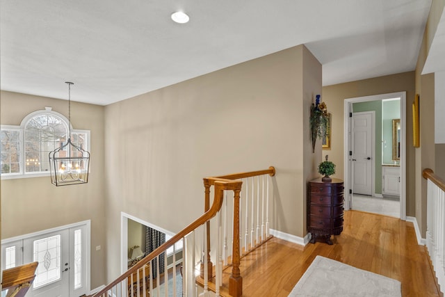 corridor featuring light wood-style floors, baseboards, a notable chandelier, and an upstairs landing