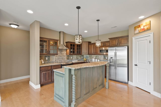 kitchen with light wood-style floors, appliances with stainless steel finishes, wall chimney range hood, light stone countertops, and tasteful backsplash