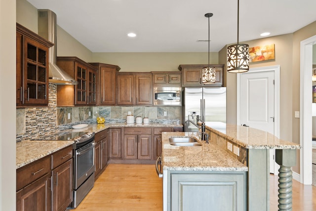 kitchen with wall chimney exhaust hood, a kitchen island with sink, stainless steel appliances, light wood-type flooring, and a sink