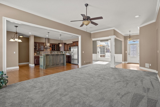 unfurnished living room featuring light colored carpet, crown molding, baseboards, and ceiling fan with notable chandelier