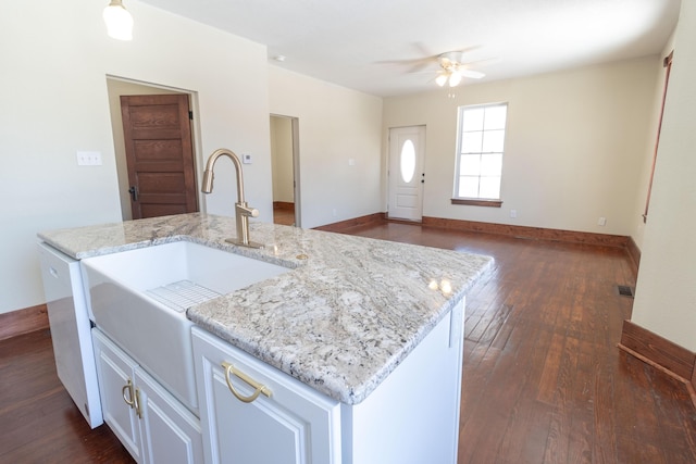 kitchen featuring a sink, baseboards, light stone countertops, dark wood finished floors, and a center island with sink