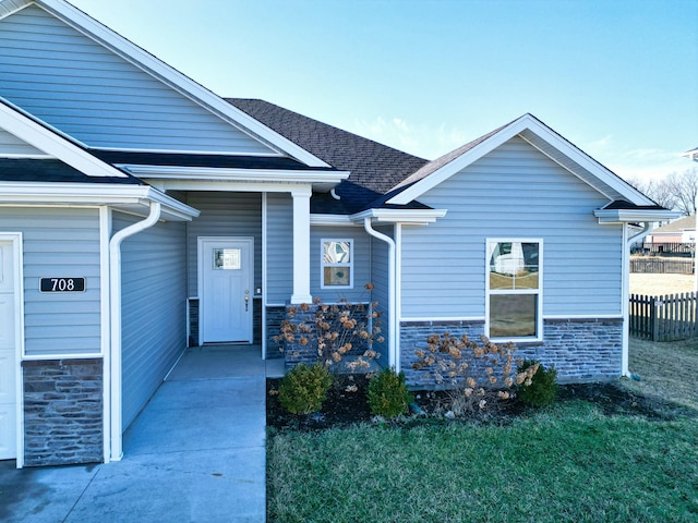 property entrance with stone siding, roof with shingles, a lawn, and fence