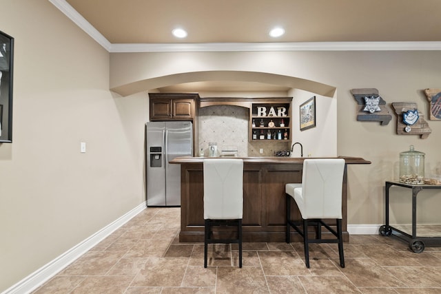 bar featuring baseboards, stainless steel refrigerator with ice dispenser, indoor wet bar, and crown molding