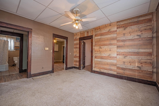 carpeted empty room featuring a ceiling fan, a drop ceiling, wooden walls, and baseboards