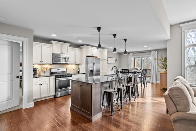 kitchen featuring a kitchen island with sink, open floor plan, hanging light fixtures, appliances with stainless steel finishes, and dark countertops