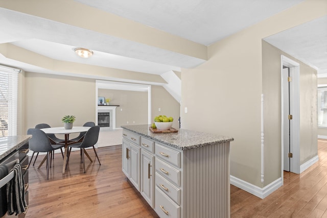 kitchen with light wood finished floors, baseboards, a glass covered fireplace, a kitchen island, and light stone countertops