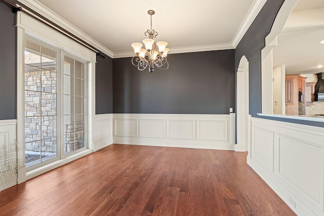 unfurnished dining area with arched walkways, a wainscoted wall, crown molding, an inviting chandelier, and wood finished floors