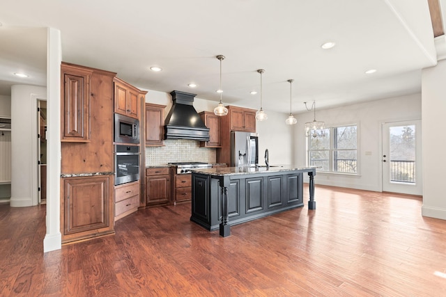 kitchen with a kitchen island with sink, premium range hood, appliances with stainless steel finishes, decorative backsplash, and dark wood-style floors