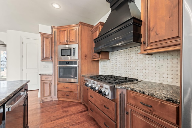 kitchen with decorative backsplash, dark stone counters, dark wood-style flooring, stainless steel appliances, and premium range hood