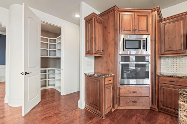 kitchen featuring appliances with stainless steel finishes, brown cabinets, dark wood-type flooring, stone counters, and backsplash