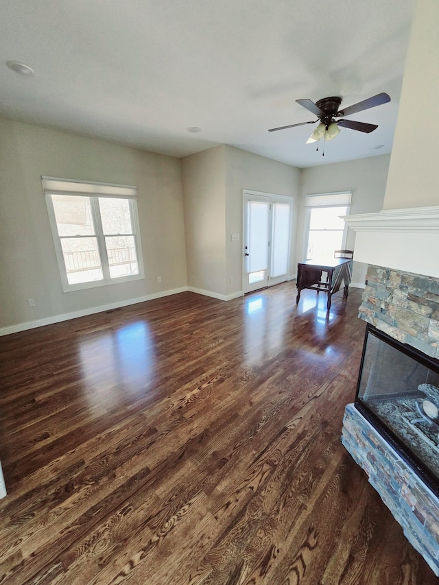 living room featuring a stone fireplace, dark wood-type flooring, a ceiling fan, and baseboards