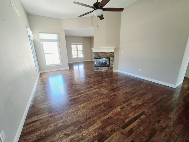 unfurnished living room featuring dark wood-style floors, a stone fireplace, a ceiling fan, and baseboards