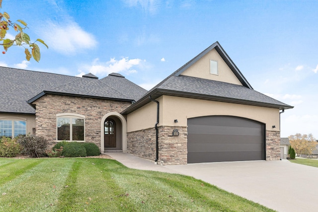 french country inspired facade with an attached garage, concrete driveway, roof with shingles, stucco siding, and a front yard