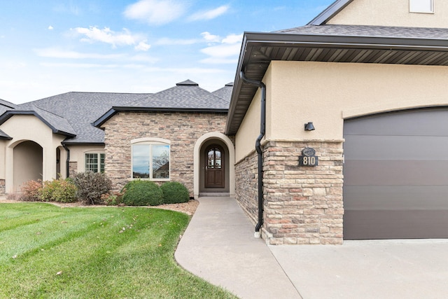 doorway to property featuring a garage, a shingled roof, stone siding, a yard, and stucco siding