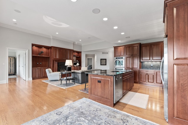 kitchen with stainless steel appliances, light wood-style floors, open floor plan, dark stone countertops, and an island with sink