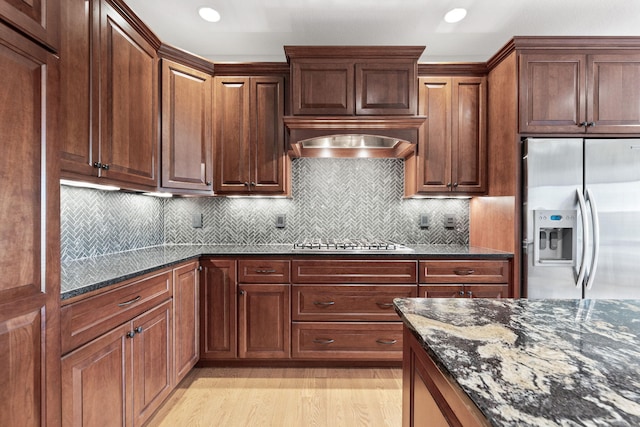 kitchen featuring light wood-style flooring, appliances with stainless steel finishes, dark stone countertops, range hood, and backsplash