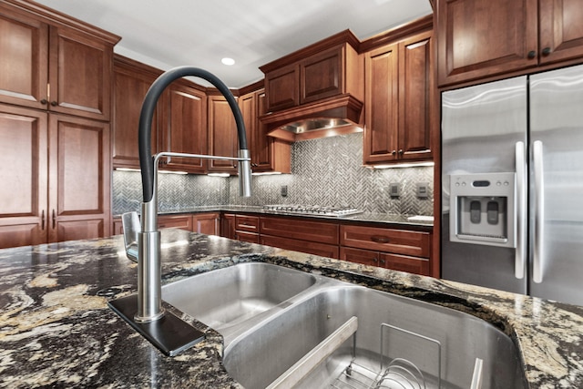 kitchen with gas cooktop, premium range hood, stainless steel refrigerator with ice dispenser, backsplash, and dark stone counters