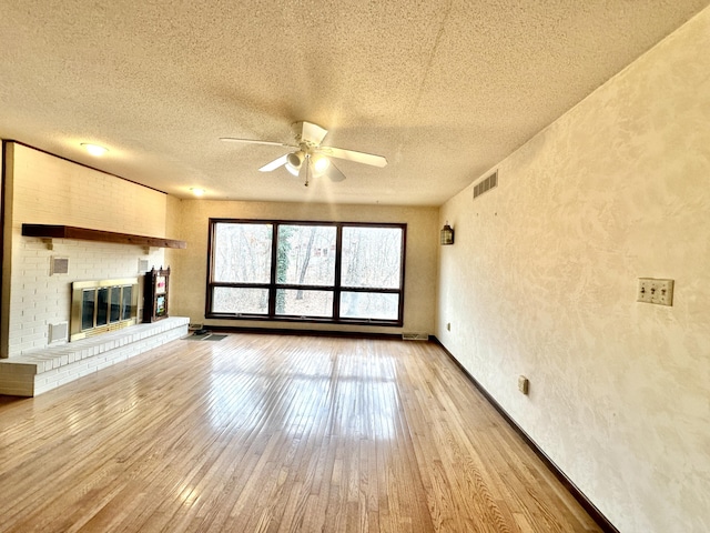 unfurnished living room with light wood-type flooring, a textured ceiling, a brick fireplace, ceiling fan, and a textured wall