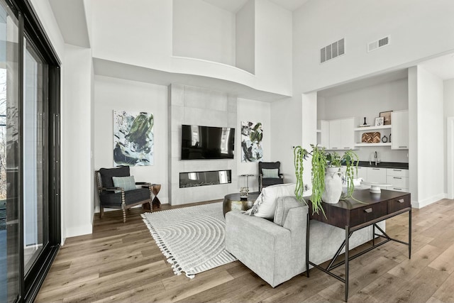 living room with light wood-type flooring, a towering ceiling, visible vents, and a glass covered fireplace