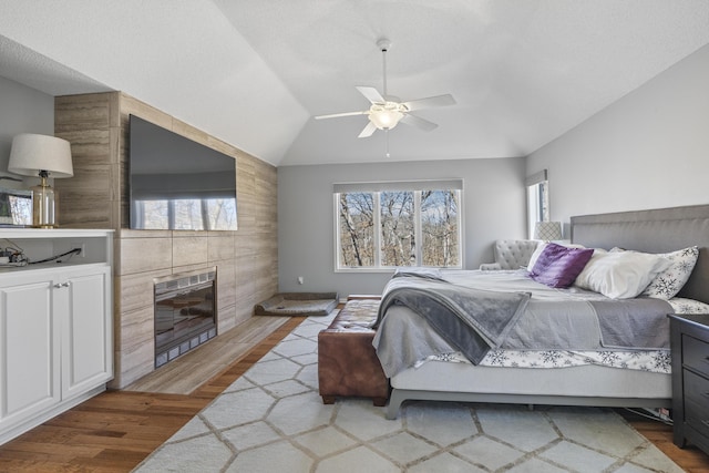 bedroom with ceiling fan, vaulted ceiling, a tiled fireplace, and wood finished floors