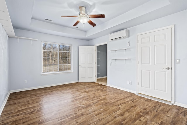 unfurnished bedroom with a raised ceiling, a wall mounted air conditioner, baseboards, and wood finished floors