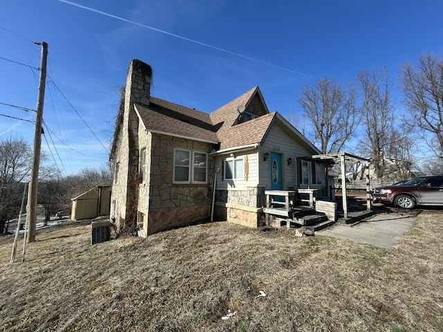 view of side of home featuring roof with shingles, a chimney, central air condition unit, stone siding, and a wooden deck