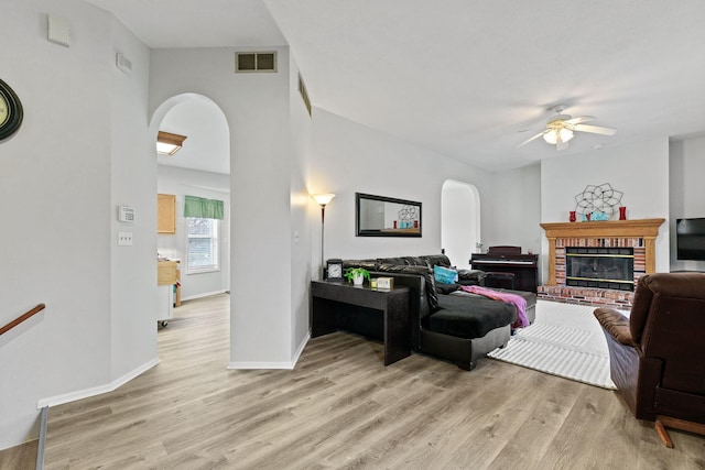 living room featuring arched walkways, visible vents, a ceiling fan, a brick fireplace, and light wood finished floors