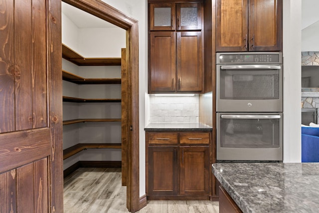 kitchen featuring decorative backsplash, dark stone countertops, light wood finished floors, and stainless steel double oven