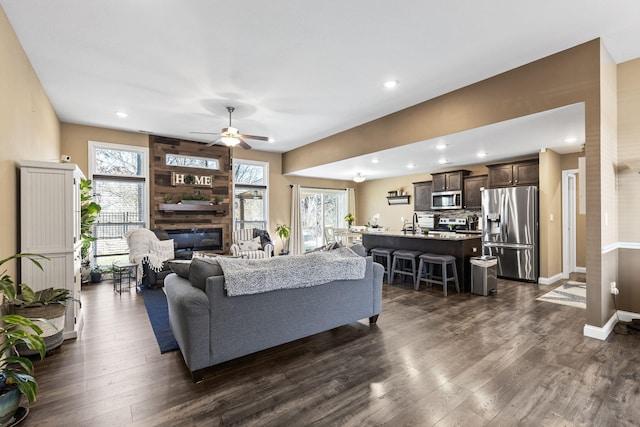 living area featuring a large fireplace, baseboards, ceiling fan, dark wood finished floors, and recessed lighting