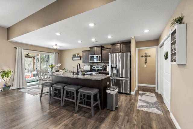 kitchen with a breakfast bar, a kitchen island with sink, dark wood-style floors, dark brown cabinetry, and appliances with stainless steel finishes