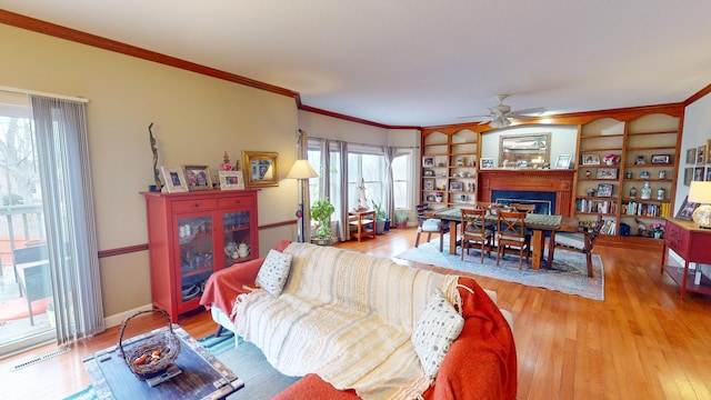 living area featuring wood finished floors, a ceiling fan, baseboards, ornamental molding, and a glass covered fireplace