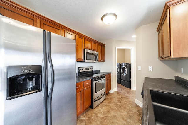 kitchen featuring brown cabinetry, light tile patterned flooring, stainless steel appliances, washer and dryer, and dark countertops