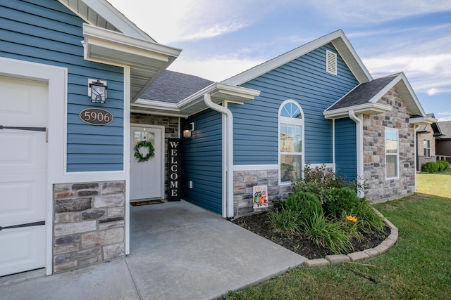 doorway to property featuring stone siding, a lawn, an attached garage, and a shingled roof