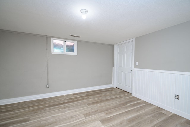 basement featuring visible vents, a wainscoted wall, a textured ceiling, light wood-style floors, and baseboards