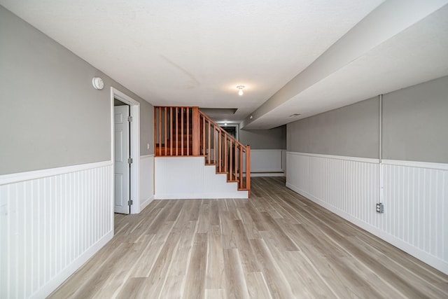 finished basement featuring stairway, light wood-style flooring, and wainscoting