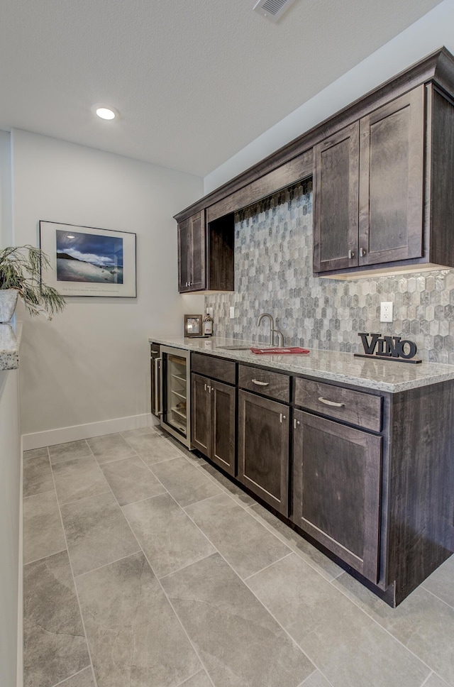 kitchen featuring backsplash, baseboards, wine cooler, dark brown cabinetry, and light stone counters