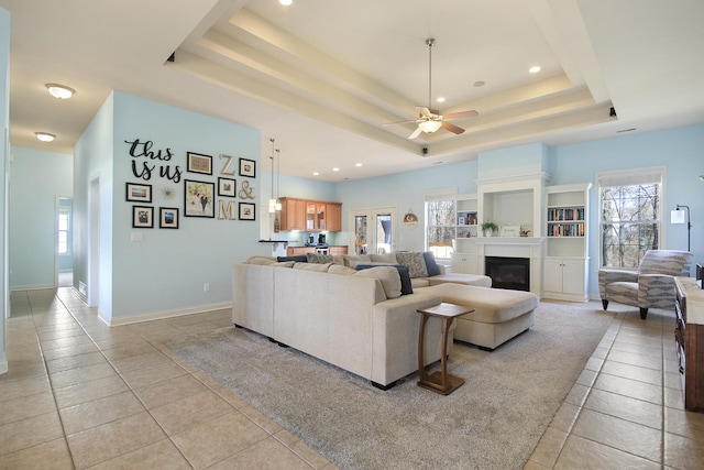 living area featuring light tile patterned floors, baseboards, ceiling fan, a glass covered fireplace, and a raised ceiling