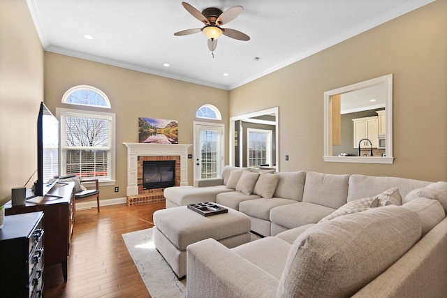 living room with wood finished floors, baseboards, ceiling fan, crown molding, and a brick fireplace