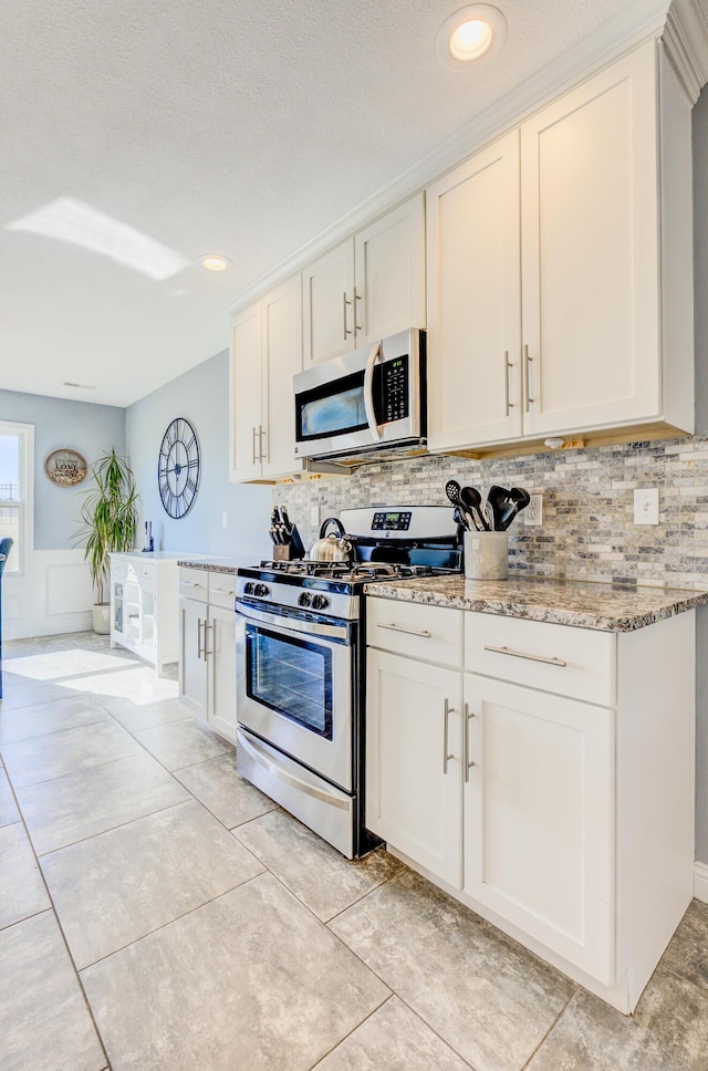 kitchen with tasteful backsplash, a wainscoted wall, light stone counters, light tile patterned floors, and appliances with stainless steel finishes